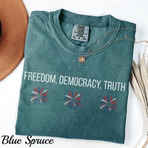 Pu&ograve; includere: T-shirt blu abete con le parole "FREEDOM, DEMOCRACY, TRUTH" in bianco. La maglietta presenta tre disegni di fuochi d'artificio in rosso, bianco e blu. Una collana d'oro con un ciondolo &egrave; drappeggiata sulla maglietta.