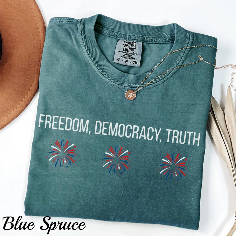 Pu&ograve; includere: T-shirt blu abete con le parole "FREEDOM, DEMOCRACY, TRUTH" in bianco. La maglietta presenta tre disegni di fuochi d'artificio in rosso, bianco e blu. Una collana d'oro con un ciondolo &egrave; drappeggiata sulla maglietta.