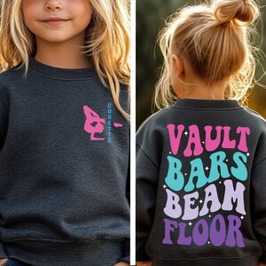 May include: Dark gray sweatshirt with a gymnastics design. The front features a pink and blue gymnast graphic and the name "Cosette". The back displays the words "Vault Bars Beam Floor" in colorful retro fonts.