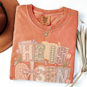 May include: Coral-colored t-shirt with the words "HE IS RISEN" in a patchwork design. The design includes floral, leopard print, and plaid patterns, along with a pink bow and butterflies. A gold necklace with a pendant is draped on the shirt.