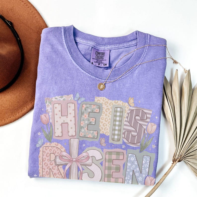 May include: A light purple t-shirt with the words "HE IS RISEN" in a patchwork design. The letters are decorated with floral, leopard print, and striped patterns. A gold necklace with a pendant is draped across the shirt.