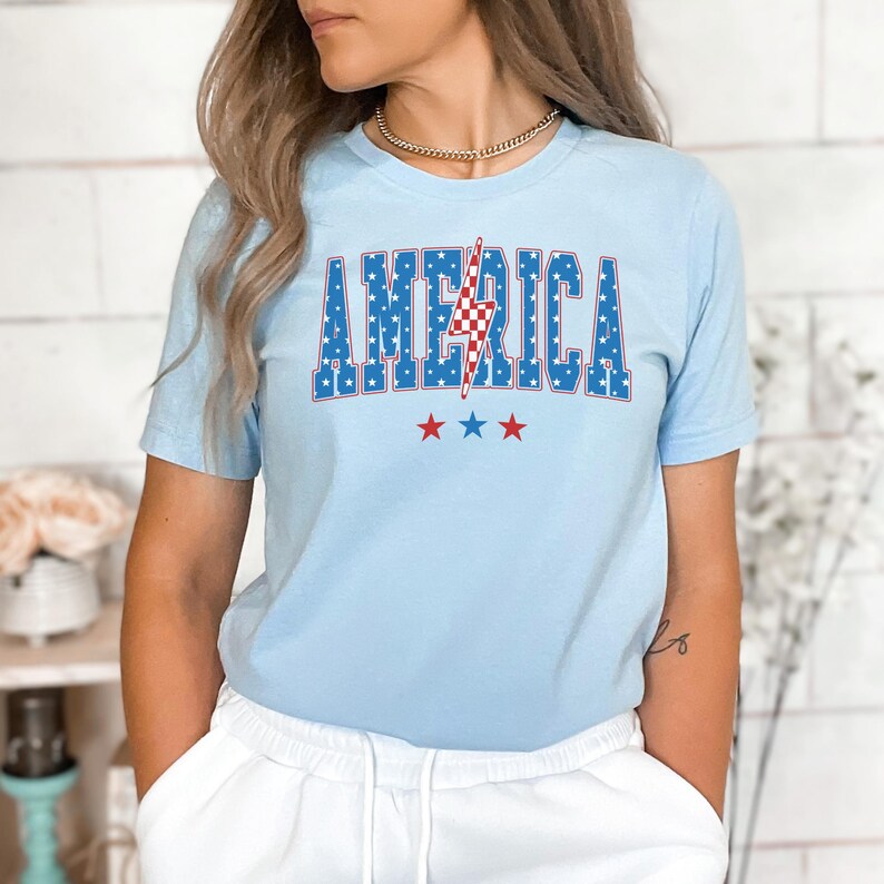 May include: Light blue t-shirt with the word "AMERICA" in blue letters with white stars. A red and white lightning bolt is in the center of the word. Three red stars are below the word.
