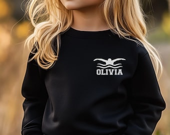Peut inclure: Sweat-shirt noir avec un motif de nageur blanc et le nom "OLIVIA" imprimé en dessous. Le sweat-shirt est de couleur noire unie et a un col rond. Le motif représente un nageur en mouvement.