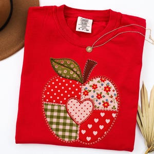 Op de afbeelding: Een felrood T-shirt met ronde hals en een grote appelvormige applicatie van verschillende stoffen. Het appelontwerp bevat rood-witte stippen, groen-wit vichy-ruitjes, bloemenpatronen en een centraal hartvormig lapje in het midden.