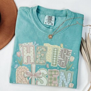 May include: A turquoise Comfort Colors t-shirt with the words "HE IS RISEN" in a patchwork design. The design includes floral, plaid, and striped patterns, with butterflies and tulips. A gold necklace with a pendant rests on the shirt.