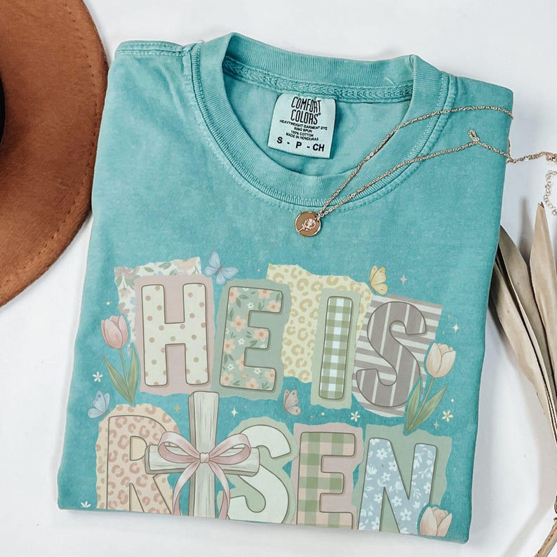May include: A turquoise Comfort Colors t-shirt with the words "HE IS RISEN" in a patchwork design. The design includes floral, plaid, and striped patterns, with butterflies and tulips. A gold necklace with a pendant rests on the shirt.