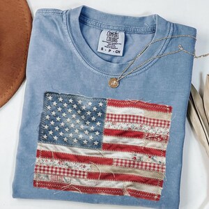 May include: A light blue Comfort Colors t-shirt with a sewn-on American flag design. The flag is made of various fabric patterns, including floral and gingham, with a distressed aesthetic. A gold necklace with a pendant rests on the shirt.