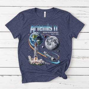 May include: Navy blue t-shirt featuring a graphic of the Artemis II mission. The design includes Earth, the Moon, a rocket launching, and a spacecraft, with the text "Artemis II Around the Moon and Back April 1, 2026."