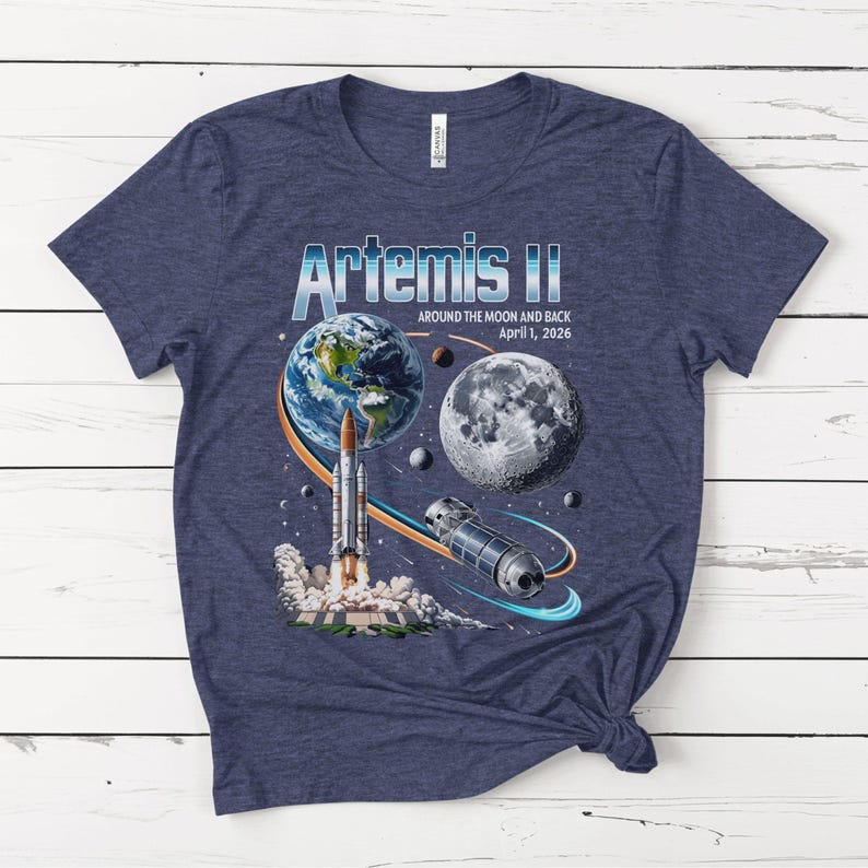 May include: Navy blue t-shirt featuring a graphic of the Artemis II mission. The design includes Earth, the Moon, a rocket launching, and a spacecraft, with the text "Artemis II Around the Moon and Back April 1, 2026."