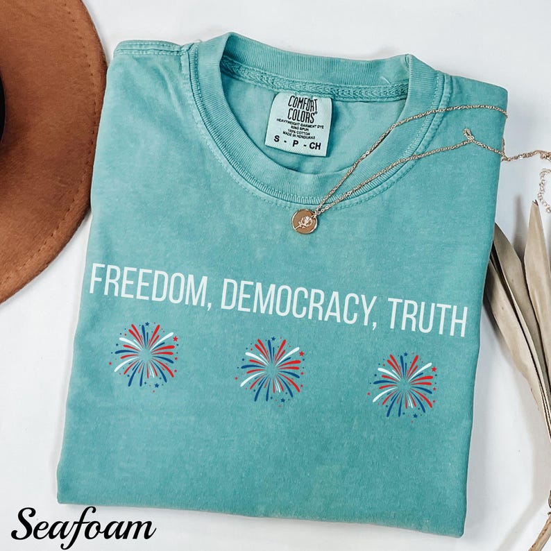 Pu&ograve; includere: T-shirt verde acqua con le parole "FREEDOM, DEMOCRACY, TRUTH" in bianco. Sotto il testo ci sono tre disegni di fuochi d'artificio rossi, bianchi e blu. Una collana d'oro con un ciondolo &egrave; sulla maglietta.