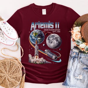 May include: Burgundy t-shirt featuring a graphic of Earth, the Moon, a rocket, and a space station. The text "Artemis II" is displayed above the graphic, with "Around the Moon and Back April 1, 2026" below. The shirt is part of a space-themed collection.
