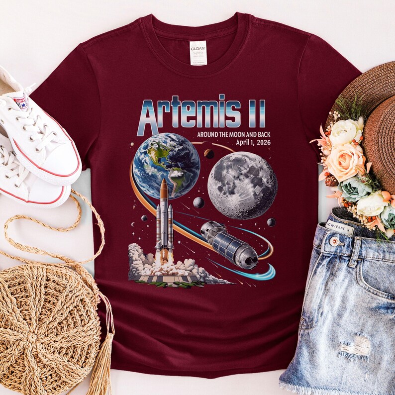 May include: Burgundy t-shirt featuring a graphic of Earth, the Moon, a rocket, and a space station. The text "Artemis II" is displayed above the graphic, with "Around the Moon and Back April 1, 2026" below. The shirt is part of a space-themed collection.