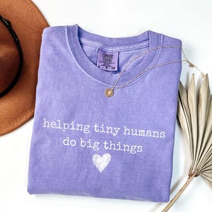 May include: A lilac t-shirt with the white text "helping tiny humans do big things" and a heart graphic. A gold necklace and a brown hat are also visible. The shirt is made of a soft material.