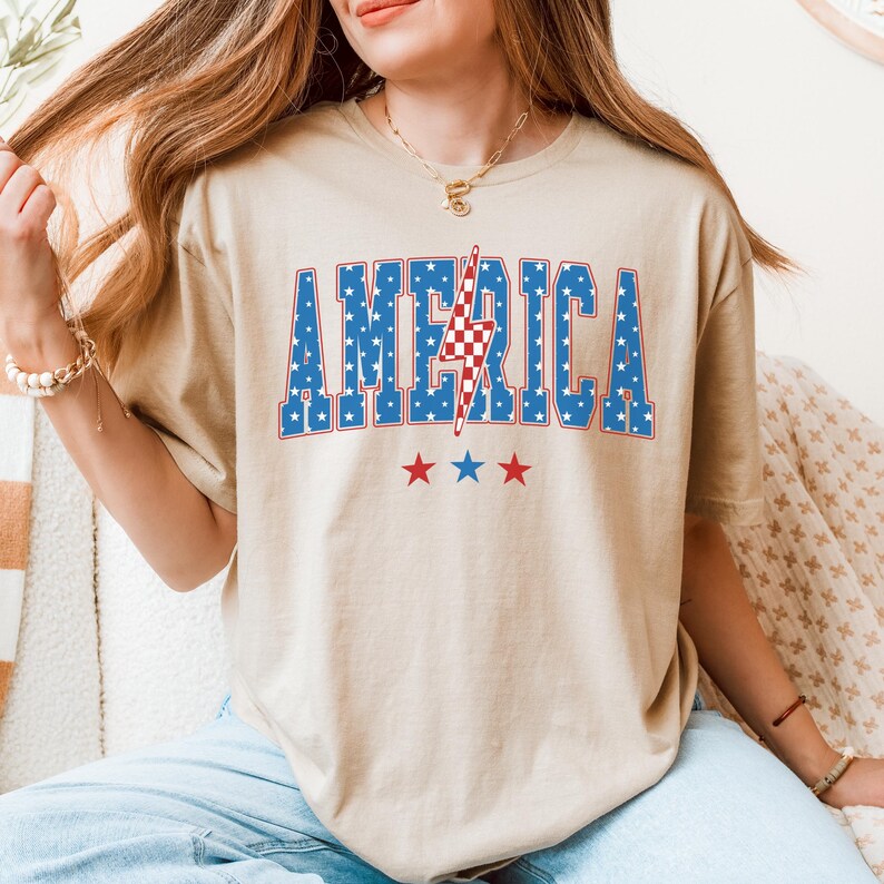 May include: Beige t-shirt with the word "AMERICA" in blue block letters filled with white stars. A red and white checkered lightning bolt is in the center of the word. Three red stars are below the word.