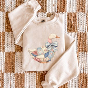 May include: A cream-colored sweatshirt featuring a whimsical goose design. The goose has a patchwork quilt-style body in shades of blue, red, and floral patterns, with a blue gingham bow. The sweatshirt has long sleeves and a crew neck.
