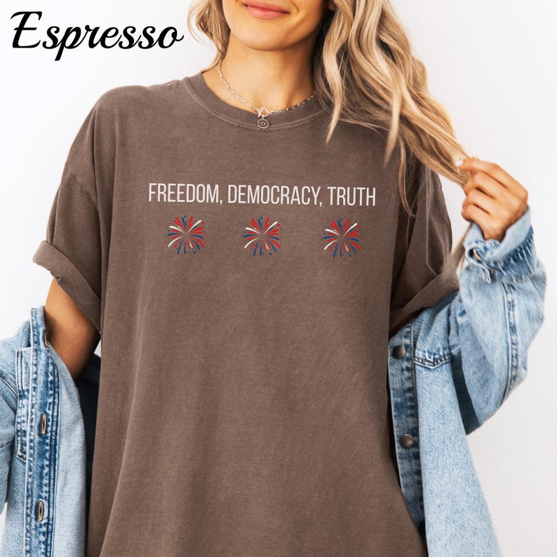 Pu&ograve; includere: T-shirt color espresso con le parole "FREEDOM, DEMOCRACY, TRUTH" in bianco. Sotto il testo ci sono tre fuochi d'artificio stilizzati in rosso, bianco e blu. La maglietta ha le maniche arrotolate e viene indossata con una giacca di jeans.