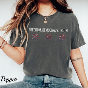 Pu&ograve; includere: T-shirt grigio scuro con le parole bianche "FREEDOM, DEMOCRACY, TRUTH". Sotto, tre disegni di fuochi d'artificio rossi, bianchi e blu. La parola "Pepper" &egrave; in basso a sinistra.