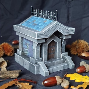 Peut inclure: Une miniature de bâtiment en pierre grise avec un toit en tuiles bleues et une clôture décorative en métal. Le bâtiment a une porte en bois et des marches. Des glands et des feuilles sont éparpillés autour de la base.
