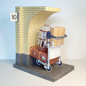 Puede incluir: Un modelo de papel de un arco de ladrillo etiquetado con el número 10, que presenta un carrito de equipaje cargado con una maleta marrón, una jaula blanca con un pequeño búho blanco y varios paquetes rectangulares de color canela.