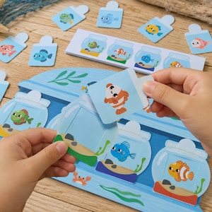 May include: A matching game featuring a blue board with illustrations of fish bowls and various colorful cartoon fish cards. The game includes a long rectangular strip with four fish bowl graphics and several individual square cards with tabs.