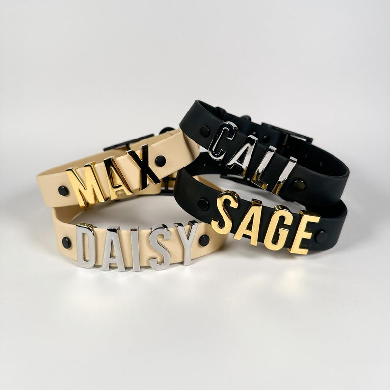 Puede incluir: Cuatro collares personalizados con nombres en cuero crema y negro. Los nombres "MAX", "DAISY", "CALL" y "SAGE" se muestran en letras met&aacute;licas doradas y plateadas. Cada collar tiene un cierre de hebilla y detalles met&aacute;licos.