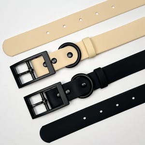 Puede incluir: Primer plano de tres cinturones en crema y negro. El cintur&oacute;n color crema tiene una hebilla negra y una anilla en D a juego. El cintur&oacute;n negro tiene una hebilla negra y una anilla en D. Los cinturones son de material liso y tienen un dise&ntilde;o minimalista.