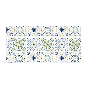 May include: White beach towel featuring a repeating pattern of blue and yellow floral designs. The pattern includes stylized flowers, stars, and geometric shapes. The towel is rectangular and appears to be made of a soft material.