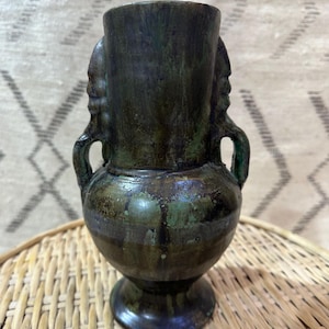 May include: A dark green ceramic vase with a bulbous body, a cylindrical top, and a pedestal base. The vase features two handles and two sculpted faces on either side. The vase is displayed on a woven wicker tray.