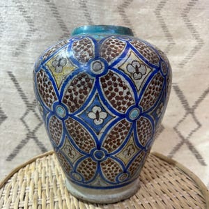 May include: A ceramic vase with a blue, brown, and white geometric pattern. The vase has a turquoise rim and a round shape. It is sitting on a woven wicker tray. The vase is a decorative item.