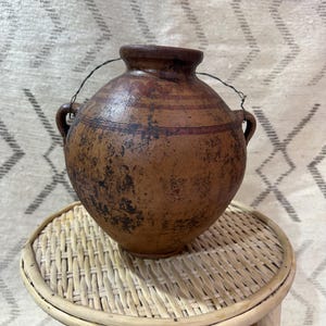 Puede incluir: Un jarrón de cerámica marrón rústico con cuerpo redondeado, dos asas pequeñas y un asa de alambre. El jarrón tiene una superficie texturizada con marcas oscuras y una banda marrón rojiza. Se encuentra sobre una pequeña mesa redonda de mimbre tejido.