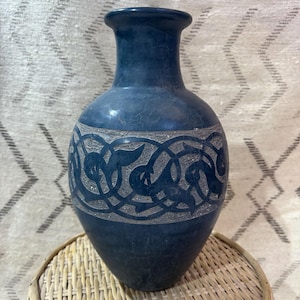 May include: A blue vase with a decorative band of carved gray patterns. The vase has a wide base, a slender neck, and a flared rim. It sits on a round, woven wicker table.