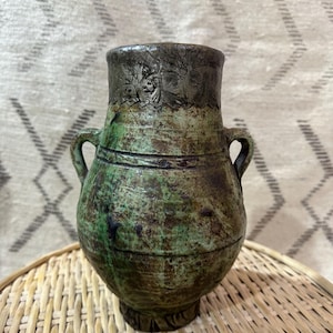 May include: A handcrafted, antique-style ceramic vase with a distressed green and brown finish. The vase features two small handles and a decorative rim. The vase is displayed on a woven wicker surface.