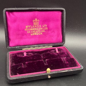 May include: An open, black jewelry box with a magenta velvet interior. The box has a gold clasp and hinges. Gold lettering inside reads "HYLANDS LTD WATCHMAKERS & JEWELLERS 32. CHEAPSIDE. LONDON."