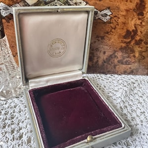 May include: An open, vintage jewelry box with a light gray exterior and a burgundy velvet interior. The box has a gold-colored clasp and trim. The inside lid has a cream-colored satin lining with a gold emblem.
