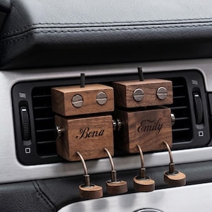 Puede incluir: Dos clips ambientadores para coche con forma de robot de madera sujetos a una rejilla de ventilación. Cada robot tiene una cabeza de madera oscura con dos ojos de tornillo plateados, un cuerpo grabado con los nombres Bona y Emily y patas de metal.