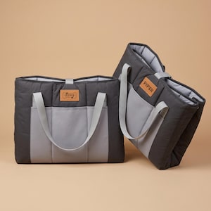 Puede incluir: Dos bolsos de mano en gris carbón y gris claro con asas dobles y bolsillos exteriores. Un bolso presenta un parche rectangular color canela con el nombre COOPER y un icono de hueso, el otro un parche color canela con el nombre PIPER.