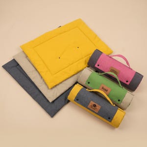 Puede incluir: Una colección de alfombrillas rectangulares para mascotas en amarillo, beige y gris oscuro. Tres alfombrillas están extendidas, mientras que tres están enrolladas con asas de transporte. Cada alfombrilla enrollada tiene un parche de cuero.