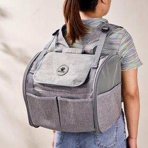 May include: A grey pet carrier backpack featuring adjustable shoulder straps, mesh side panels for ventilation, and two front pockets. A circular patch on the top flap displays a paw print icon and the text Buddy in black lettering.