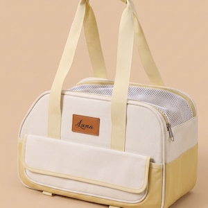 Puede incluir: Un transportín para mascotas de color crema y amarillo pálido con un parche rectangular de cuero marrón que lleva la palabra Luna impresa en cursiva negra. Incluye dos asas superiores, un bolsillo frontal y una malla blanca de ventilación.