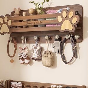 Peut inclure: Un organisateur mural en bois foncé avec deux grands accents latéraux en forme de patte et le texte "Rosie's holder" gravé sur la barre avant. L'unité comprend une étagère supérieure et sept crochets métalliques, chacun orné d'une patte.