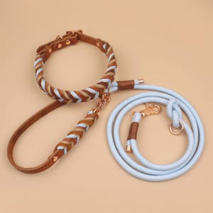 May include: A braided leather dog collar and leash set featuring tan, brown, and light blue strands with rose gold hardware. The collar has a buckle closure, and the matching light blue rope leash includes a rose gold swivel clasp and metal accents.
