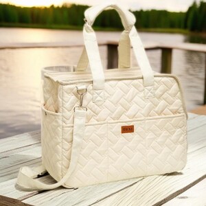 May include: A white quilted tote bag with a long shoulder strap and a zippered closure. The bag has a small pocket on the front with the word "DEMI" printed on it.