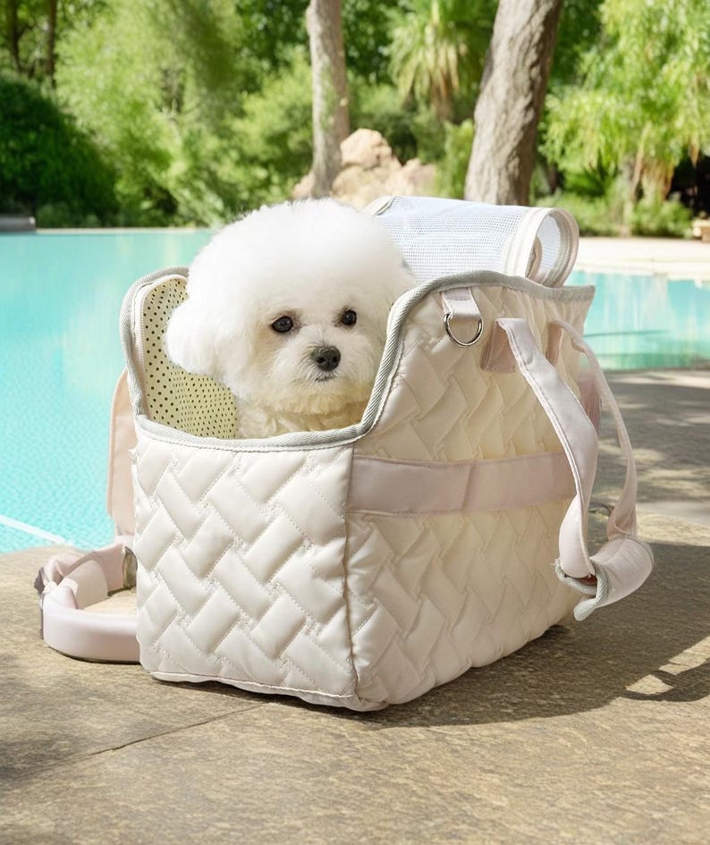 May include: A white quilted pet carrier with a white dog inside. The carrier has a pink strap and a mesh window. The carrier is sitting on a concrete surface near a pool.