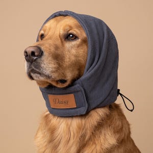 May include: A dark gray ribbed fabric snood worn by a golden-colored dog. The head covering features a rectangular tan leather-like patch on the front with the name Daisy printed in a dark script font and an adjustable black drawstring cord at the back.