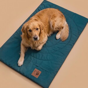Könnte beinhalten: Eine rechteckige, blaugrüne Haustiermatte mit einem gesteppten kreisförmigen Muster und einem braunen, lederartigen Aufnäher mit Pfotenabdruck-Logo. Ein goldfarbener Hund liegt auf der flach ausgelegten Matte mit glatter Stoffoberfläche.