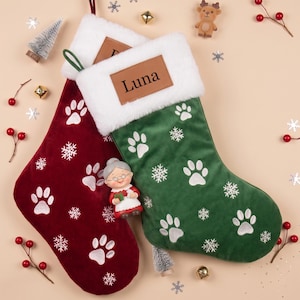 Può includere: Un paio di calze natalizie in velluto, una rosso scuro e una verde bosco, entrambe con risvolti in pelliccia sintetica bianca. La calza verde presenta una targhetta in pelle marrone con il nome Luna stampato in nero, decorata con zampe e fiocchi.