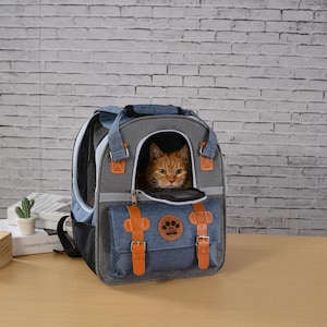 以下が含まれることがあります： オレンジ色の猫が入った、グレーとブルーのペットキャリーバックパック。 キャリアには、茶色の革ストラップと足跡のデザインが付いたデニムのフロントポケットが付いています。 バックパックにはハンドルとメッシュのサイドパネルが付いています。