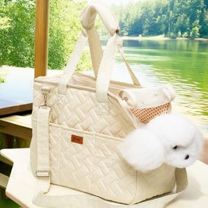 May include: A cream-colored quilted pet carrier with a zippered top, a mesh window, and a removable shoulder strap. The carrier has a small pocket on the front with the word "Baby" printed on it. A white fluffy dog toy is partially inside the carrier.