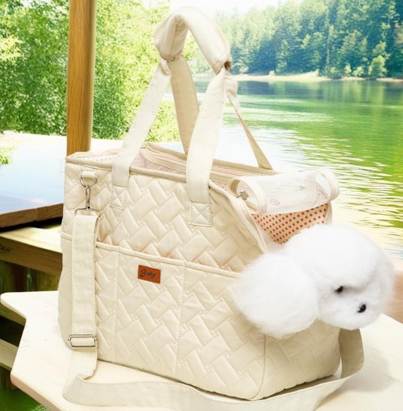 May include: A cream-colored quilted pet carrier with a zippered top, a mesh window, and a removable shoulder strap. The carrier has a small pocket on the front with the word "Baby" printed on it. A white fluffy dog toy is partially inside the carrier.