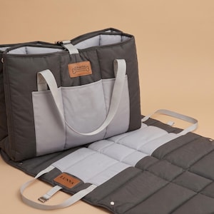 Puede incluir: Un bolso transportín acolchado para mascotas en gris oscuro y gris claro con una alfombrilla desplegable. El bolso cuenta con dos asas gris claro, un cierre de hebilla superior y dos parches de cuero sintético color canela con Charlie y LUNNA.